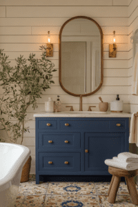 A Mediterranean-inspired bathroom featuring a navy blue vanity with gold hardware, a white marble countertop, and an arched gold-framed mirror. The shiplap walls are painted white, adding a clean and airy feel to the space. On the left, an olive tree adds a touch of greenery, while patterned blue and beige tile flooring enhances the old-world charm. Warm lighting from vintage-style sconces on either side of the mirror completes the elegant and cozy ambiance. A small wooden stool with folded white towels is placed to the right of the vanity, adding a rustic touch.