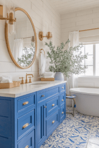 A Mediterranean-inspired bathroom featuring a bold royal blue vanity with gold hardware and a round gold-framed mirror. The walls are adorned with white shiplap, complemented by a large vase of greenery. A freestanding white bathtub and blue-and-white patterned floor tiles complete the design, adding a vibrant yet elegant touch to the space.